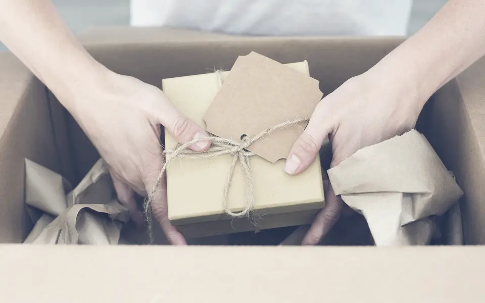 Two hands placing a wrapped gift with a kraft-paper tag into a shipping box